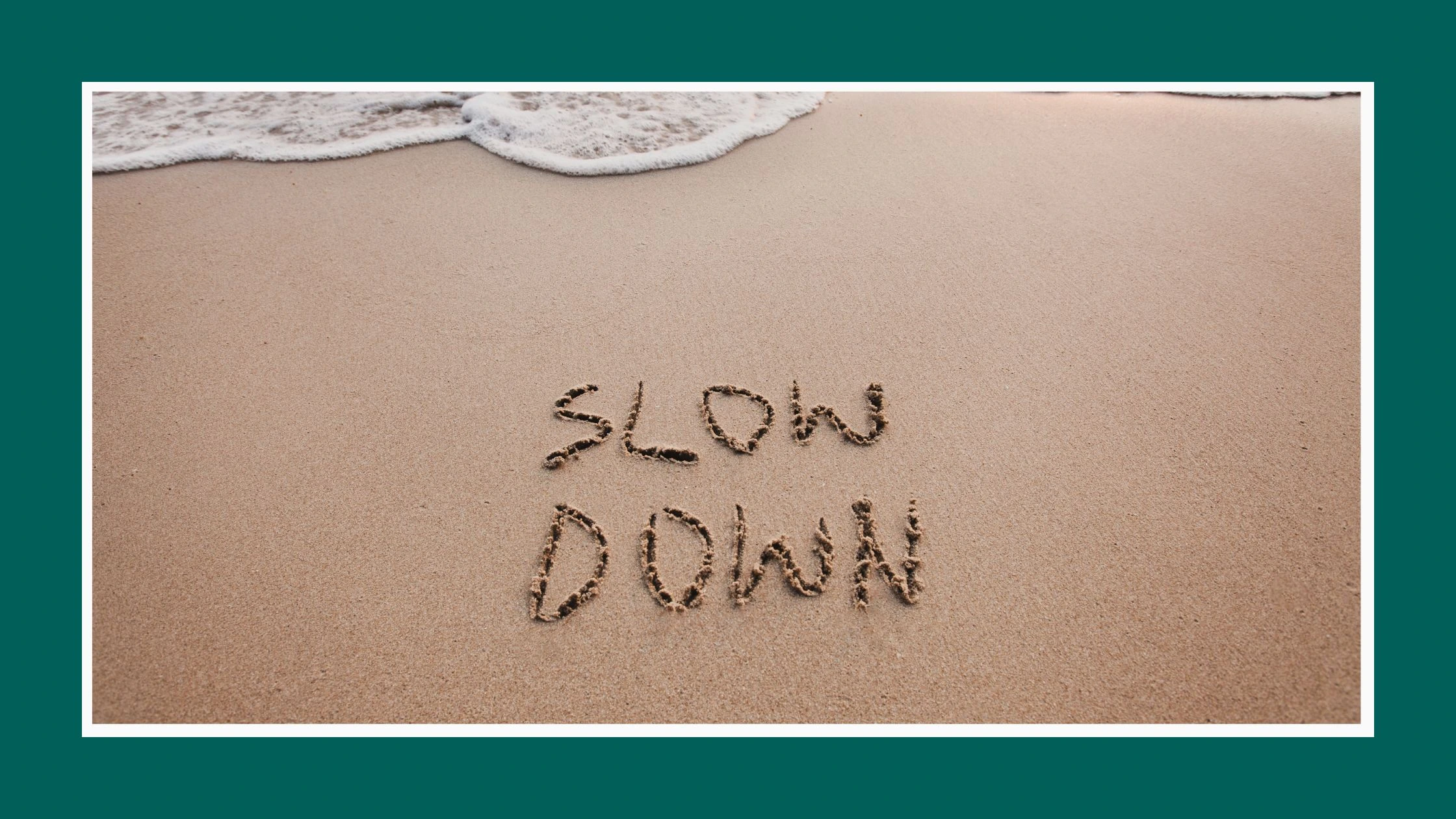 Spokojna plaża i napisane na pisaku palcem albo patykiem: Slow down. Zdjęcie wprowadzające do artykułu Uważność: jak zacząć być „tu i teraz”
